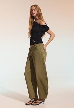 Frau mit langem blondem Haar, die ein schwarzes schulterfreies Oberteil, lockere olivgrüne Hosen und schwarze Sandalen mit Absatz trägt, seitlich stehend.