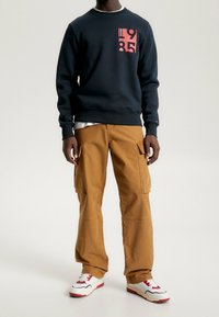 Hombre vestido con sudadera navy con estampado rojo "1985", pantalones cargo marrones y zapatillas blancas con acentos rojos, de pie sobre un fondo blanco.