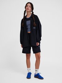 Jonge vrouw met gevlochten haar, die een zwarte oversized jas, zwarte shorts, een blauw crop top, witte sokken en blauwe sneakers draagt, staand op een witte achtergrond.