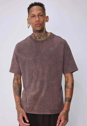 Homme avec des tatouages sur le cou et les bras porte un t-shirt marron foncé délavé à manches courtes et un pantalon noir sur un fond clair uni.
