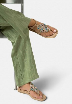 Feet wearing beige sandals decorated with multicolored gems, crossed under green textured pants while sitting on a chair.