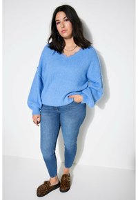 Pull en tricot texturé bleu clair avec un col en V, associé à un jean bleu ajusté et des sabots à imprimé léopard. Le mannequin se tient devant un fond blanc.