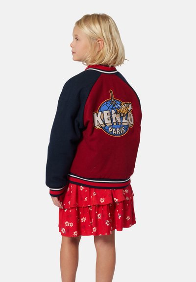 Rode en marineblauwe varsity jack met een ronde KENZO Paris-logo en grafisch ontwerp op de rug, voorzien van geribbelde manchetten en onderzoom, gedragen over een rode bloemenjurk.
