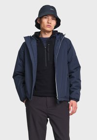 Jeune homme portant un chapeau bob bleu marine, une veste légère bleu marine par-dessus un pull zippé noir, et un pantalon foncé, debout les mains dans les poches.