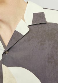 Cămașă cu mâneci scurte dintr-un material neted, având modele geometrice mari în nuanțe de gri și alb murdar, cu guler pliat și detalii cu nasturi.