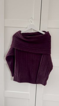 Fioletowy prążkowany sweter z odkrytymi ramionami. Posiada długie rękawy i teksturowaną, miękką tkaninę. Zawieszony na białym wieszaku.