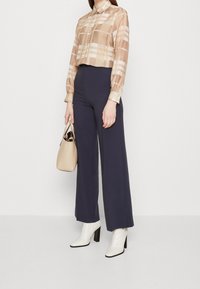 Beige ternet button-up skjorte, navy brede bukser, hvide ankelstøvler med blokhæle, og en lille beige håndtaske.