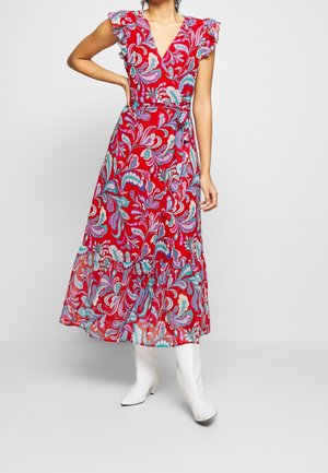 Žena v červených květovaných zavinovacích šatech s volánkovými rukávy a lemem, v kombinaci s bílými polokolenními botami, stojící na jednobarevném pozadí.