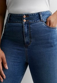 Vaqueros de denim azul de cintura alta con una textura suave, que presentan dos botones de cobre y bolsillos laterales. Las costuras contrastan en un tono más oscuro.