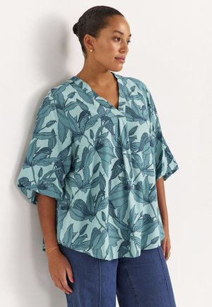 Vrouw met haar in een knot, gekleed in een blauw bloemetjespatroon blouse met wijde mouwen en een blauwe spijkerbroek, staand tegen een witte muur.