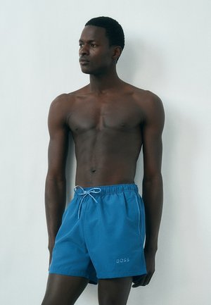 Hemdloser Mann steht an einer weißen Wand und trägt blaue Shorts mit Kordelzug und "BOSS"-Logo am linken Bein.