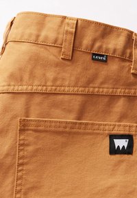 Oranžová kanvasová tkanina Levi's s zadní kapsou a černou etiketou. Obsahuje kontrastní švy a hladkou texturu s designem obdélníkového záplatování.