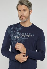 Granatowa, długoramienna koszulka wykonana z miękkiego materiału. Posiada nadrukowany tekst i grafikę na górnej lewej stronie; okrągły dekolt i wydłużone mankiety.