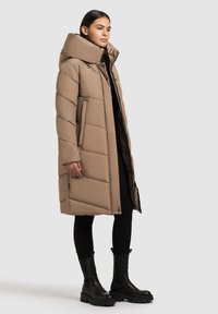 Kvinde iført en lang beige dunjakke med høj krave, sorte leggings og kraftige sorte støvler, stående imod en ensfarvet lys baggrund.