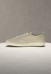 Žemo profilio smėlio spalvos sportbačiai su baltais batraiščiais, tekstūruotu padu ir kilpa ant neutralios pilkos spalvos fono.