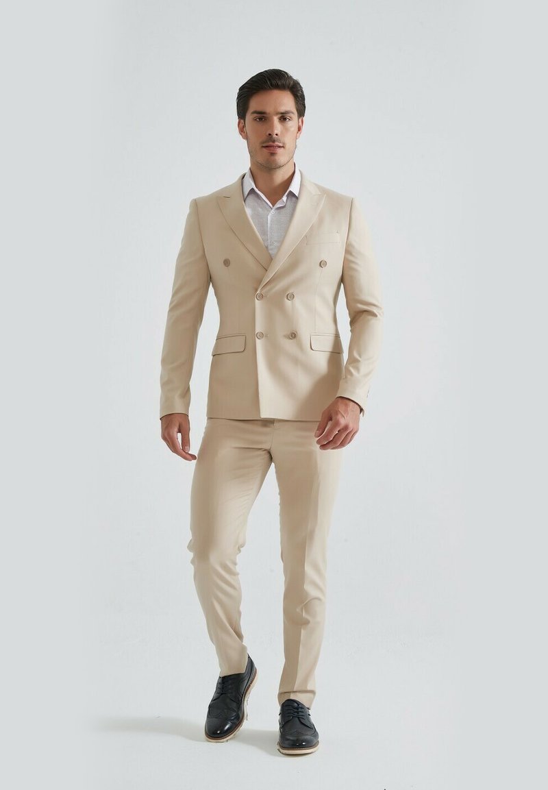 Costume beige à double boutonnage avec une coupe slim, associé à une chemise claire et des chaussures noires. Tissu lisse, lignes épurées et design ajusté.