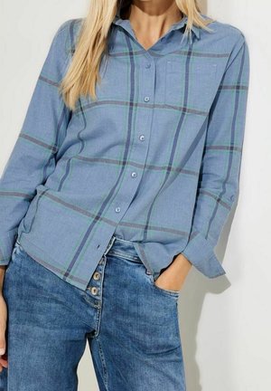 Kobieta ubrana w niebieską koszulę w kratę zapinaną na guziki, lekko wpuszczoną w niebieskie, wysokie spodnie dżinsowe z widocznymi guzikami, ręka w kieszeni.