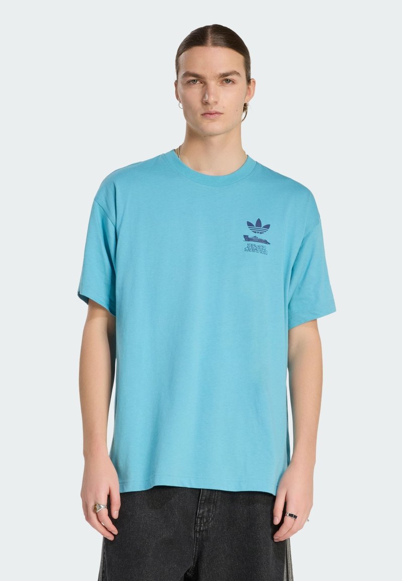 Giovane con capelli lunghi che indossa una maglietta blu Adidas con un logo scuro, pantaloni neri e anelli d'argento su entrambe le mani.