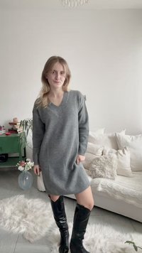 Νεαρή γυναίκα με γκρι φόρεμα από πουλόβερ και μαύρες μπότες μέχρι το γόνατο, στέκεται σε ένα φωτεινό σαλόνι με λευκό καναπέ και πράσινο τραπεζάκι.