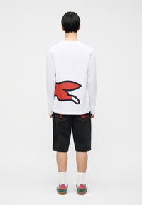 Άτομο που φοράει λευκό μακρυμάνικο πουκάμισο με κόκκινο και μαύρο σχέδιο στην πλάτη, μαύρο σορτς μέχρι το γόνατο, λευκές κάλτσες και πολύχρωμα αθλητικά παπούτσια.