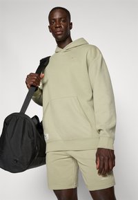 Sweatshirt à capuche vert olive clair avec épaules texturées et poche kangourou, associé à un short assorti. Sac de sport noir à proximité.