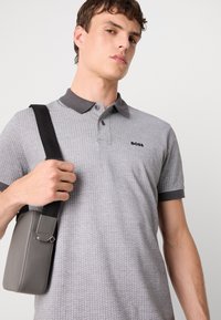 Γκρι πόλο πουκάμισο με σχέδιο, σκούρο γκρι γιακά και λογότυπο. Μοντέλο κρατάει μία μικρή γκρι τσάντα με ιμάντα ώμου.