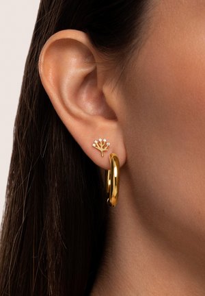 Goldene Creolen und kleine baumförmige Ohrstecker mit glitzernden Akzenten, vor einem glatten Hautton und dunklem Haar-Hintergrund.