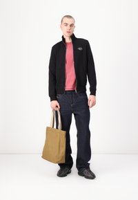 Sweatshirt preto com fecho éclair sobre uma t-shirt vermelha, combinado com jeans azuis e ténis pretos. Bolsa tote bege com alças segurada em uma mão.