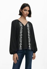 Juoda blusa su ilgomis pūstomis rankovėmis, dekoruota baltomis gėlių siuvinėjimo detalėmis V formos iškirpėje ir priekyje. Lengva medžiaga su lygiu paviršiumi.