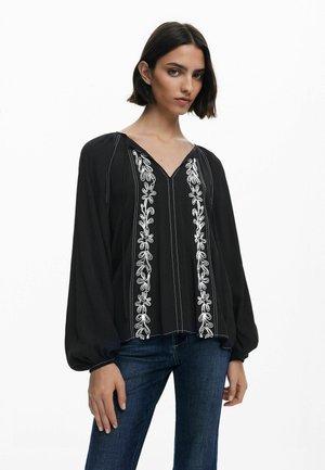 Blusa nera con maniche lunghe a sbuffo, decorata con ricami floreali bianchi lungo il collo a V e sul davanti. Tessuto leggero con una consistenza liscia.