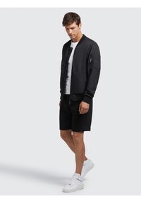 Zwarte bomberjack met een gestructureerde afwerking, rits- en drukknoopsluitingen, zijzakken en ribgebreide manchetten, gecombineerd met zwarte shorts en witte sneakers.