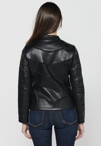 Veste en cuir noir au design ajusté, présentant des panneaux en chevron et des accents côtelés sur les manches. Portée sur un jean en denim foncé.