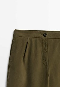 Gros plan sur un pantalon vert olive avec une ceinture à fermeture par bouton et un pli avant du côté droit.