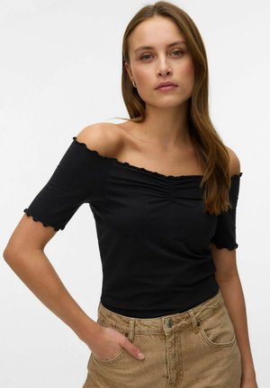 Vrouw met lang bruin haar, gekleed in een zwart off-shoulder top en beige broek, poseert met één hand in haar zak tegen een witte achtergrond.