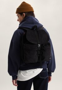 Zwarte ribfluwelen rugzak met afgeronde klep, verstelbare bandjes en twee voorzakken. Draagt wordt over een marineblauwe hoodie en een wit shirt.