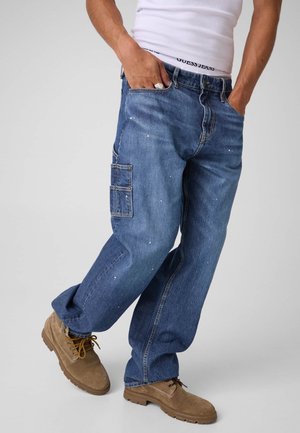 Uomo che indossa jeans cargo di denim blu larghi con piccole macchie di vernice bianca, stivali beige e una canottiera bianca con una fascia parzialmente visibile in vita.