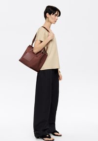 Bolso tote marrón hecho de cuero suave con dos asas cortas, un bolsillo frontal y un diseño minimalista. El modelo lleva una blusa beige y pantalones negros.