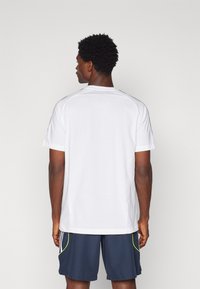 Camiseta blanca de manga corta con un cuello redondeado acanalado, hecha de un tejido suave. Las características notables incluyen acentos negros en contraste en las mangas.