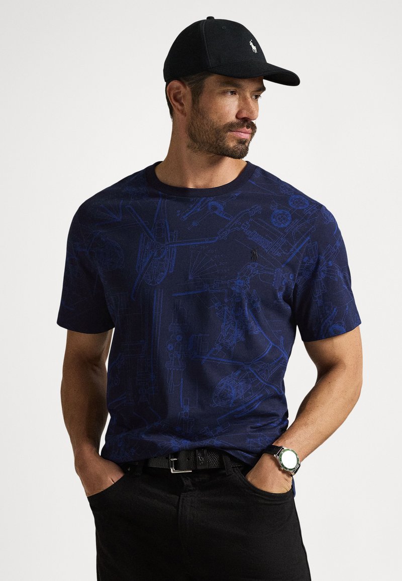 T-shirt de algodão azul-marinho com design gráfico de esquemas mecânicos, decote redondo, mangas curtas, combinado com calças pretas e um relógio preto.