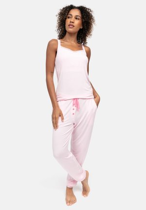 Donna con capelli ricci che indossa un top smanicato rosa chiaro e pantaloni coordinati con coulisse rosa, in piedi a piedi nudi su uno sfondo bianco.