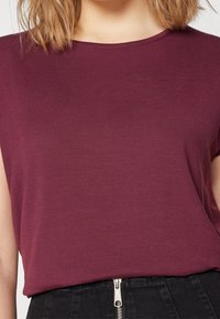 Burgundská halenka s krátkými rukávy z měkké látky, s kulatým výstřihem a volně splývavým designem, kombinovaná s černou sukní s vysokým pasem a zipem.