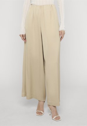Mujer con pantalones anchos beige, blusa blanca translúcida de manga larga y sandalias blancas de tacón abierto, de pie sobre un fondo liso.