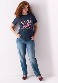 Temno modra majica s kratkimi rokavi z napisom "SANTA BABY" v rožnati in beli barvi, obogatena z barvitimi zvezdami; kombinirana s svetlo modrimi kavbojkami in rdečimi čevlji na natikanje.