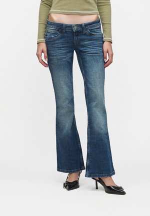 Žena oblečená v vyblednutých modrých bootcut džínsoch, čiernych špicatých vysokých podpätkoch a krátkom olivovo zelenom top s dlhým rukávom na jednoliatom pozadí.
