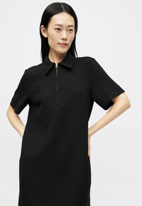 Mulher com cabelo escuro e longo, vestindo um vestido preto de mangas curtas com colarinho e fecho éclair à frente, de pé com as mãos na cintura contra um fundo branco.