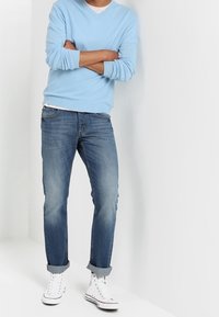 Person som står med armarna i kors, iklädd en ljusblå tröja, uppvikta blå jeans och vita högskor, framför en vit bakgrund.