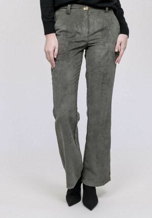 Femme portant un pantalon en velours côtelé vert olive taille haute et des bottines noires à bouts pointus, debout les mains détendues le long du corps.