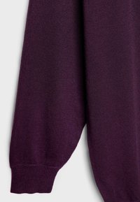 Gros plan d'une manche longue avec poignet côtelé en tissu tricoté violet foncé suspendue sur un fond clair.