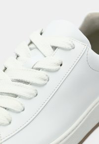 Άσπρο αθλητικό παπούτσι από λείο δέρμα με επίπεδα άσπρα κορδόνια, ενισχυμένη μύτη και λαστιχένια σόλα με διακριτικές λεπτομέρειες υφής.