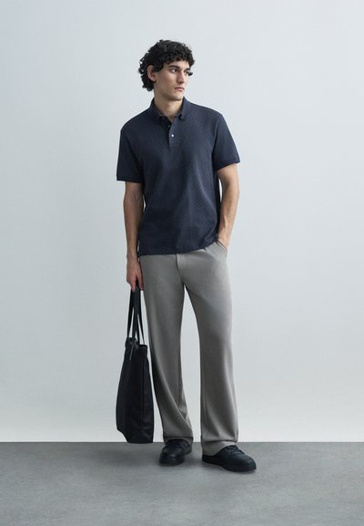 Tumši zils polo krekls ar teksturētu rakstu, īsām piedurknēm, divām pogām, kopā ar gaiši pelēkām biksēm ar krokām un melniem sporta apaviem, tur melnu somu.